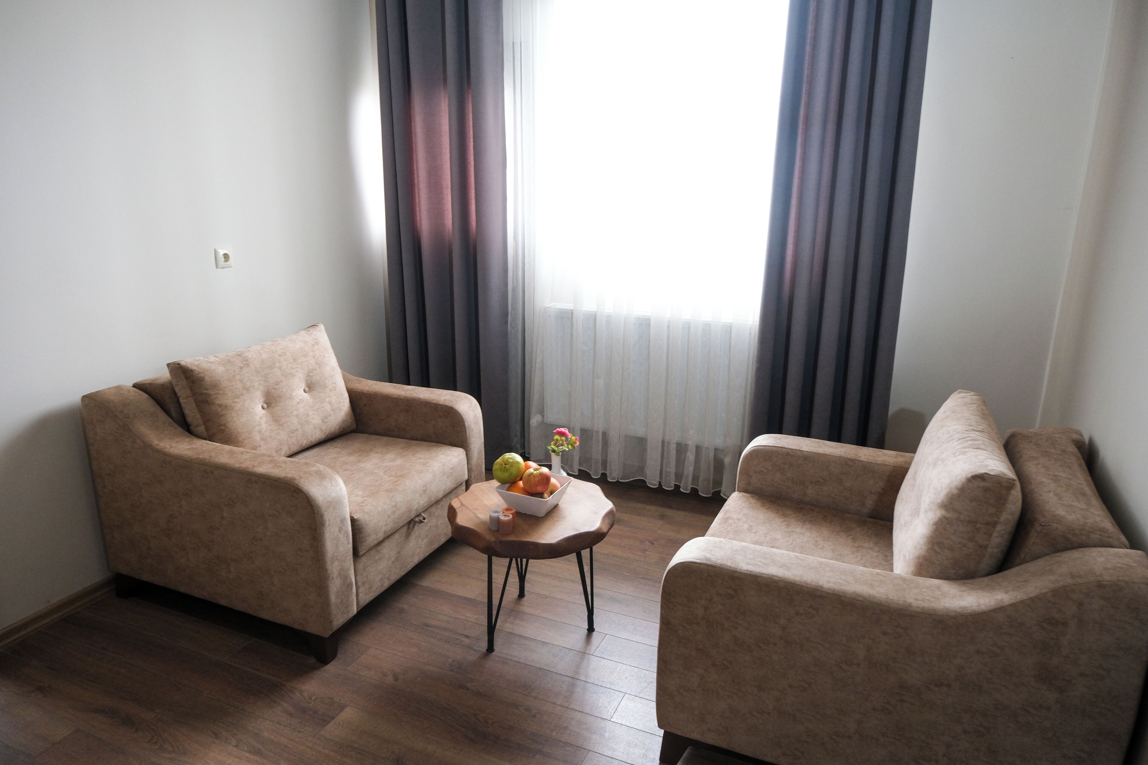 Un salon avec deux canapés beige, une petite table basse en bois sur laquelle sont disposés des fruits et un vase, et une grande fenêtre avec des rideaux.