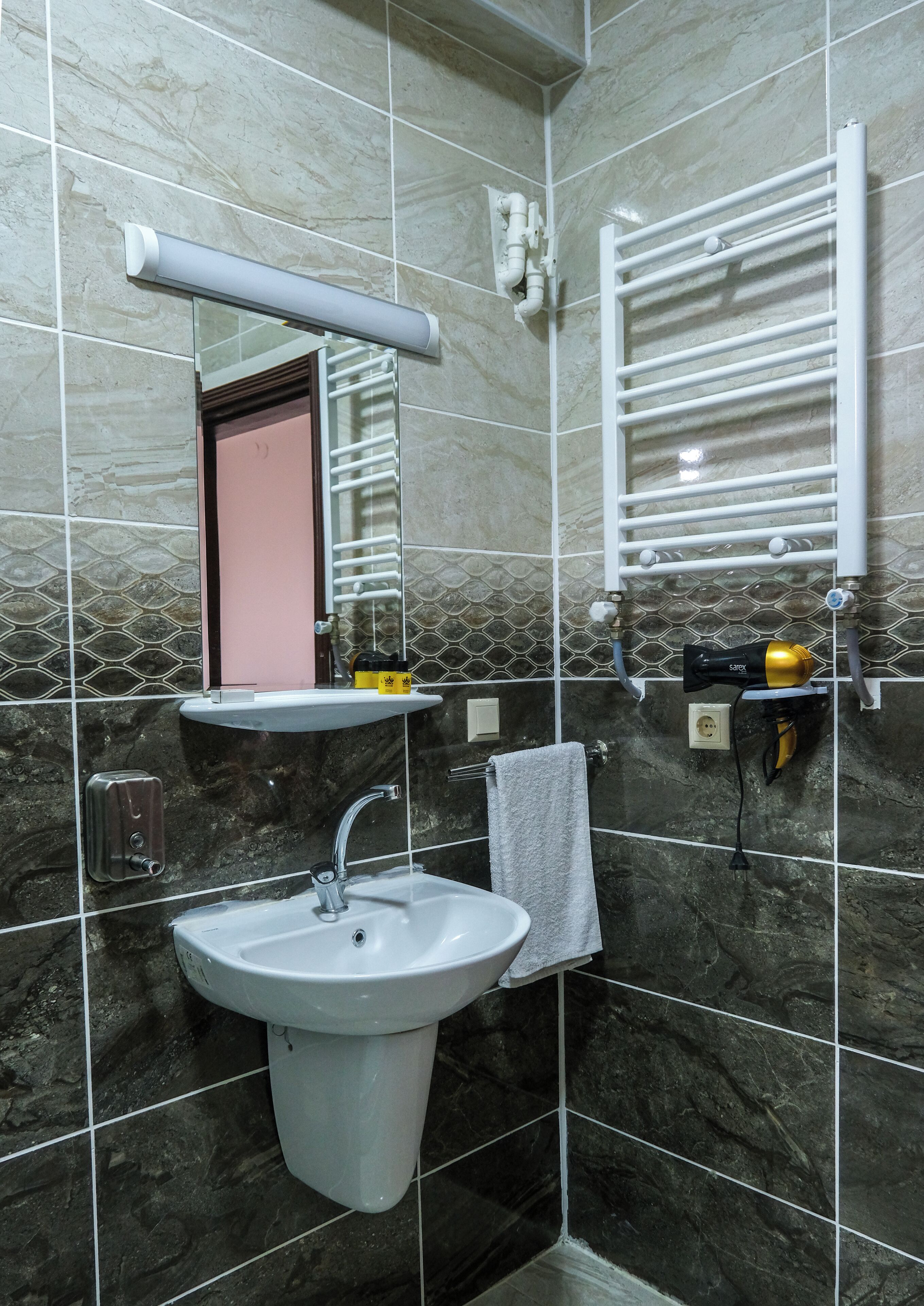 A bathroom with a pedestal sink, a towel hanging on the wall, a mirror, and a towel rack.