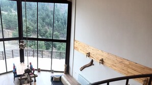 A spacious living area with a large window overlooking a forested area, a wooden beam wall, and a staircase leading to an upper level. - Savsat Kosk Otel (Savsat)