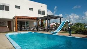 Une piscine avec un toboggan, un espace extérieur aménagé pour s’asseoir et une terrasse couverte.