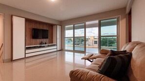 Un salon spacieux doté d’une grande fenêtre, d’un téléviseur à écran plat fixé sur un meuble en bois et d’un canapé beige agrémenté de coussins.