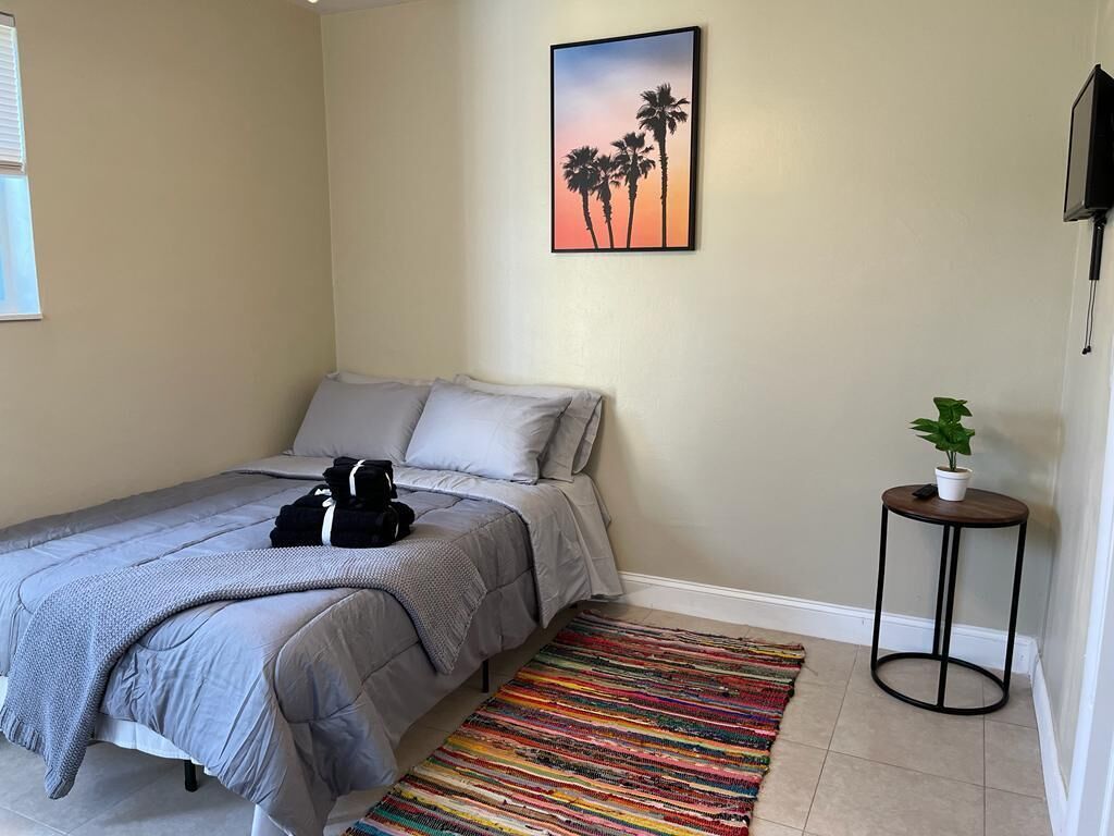 Une chambre à coucher avec un lit, des oreillers, un tapis coloré, un téléviseur fixé au mur et une petite table sur laquelle est posée une plante.