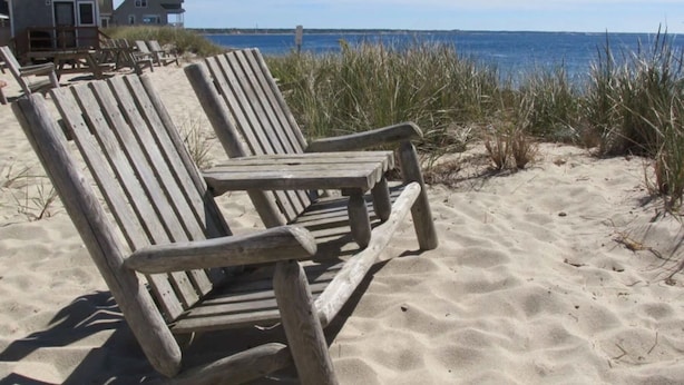 Two wooden beach chairs on a sandy beach with a view of the ocean and houses in the background.