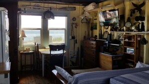 A compact room with a bed, a small table, and a chair. There is a window providing a view of the beach.