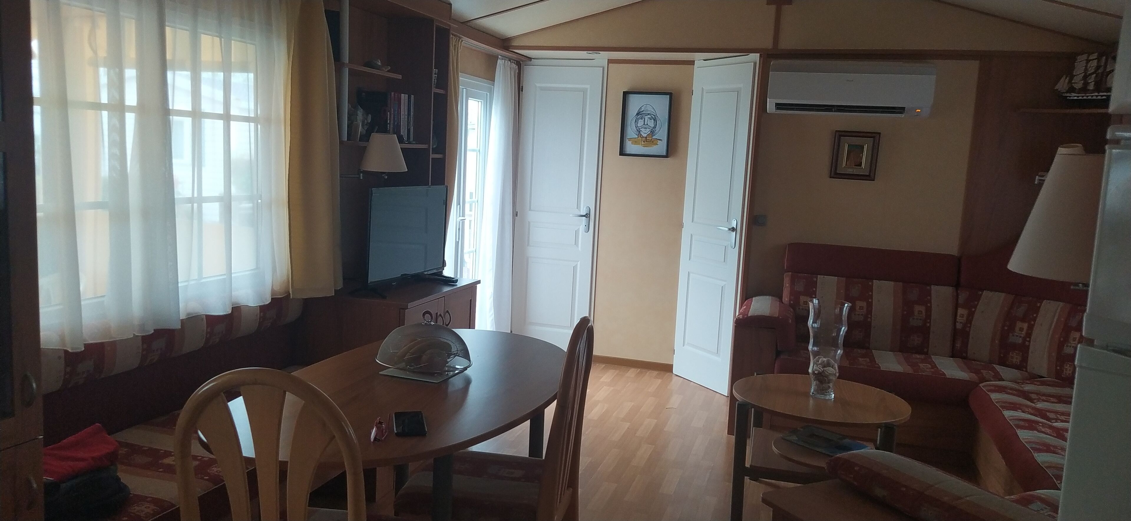 A cozy living room with a wooden table, chairs, a sofa, and a TV. There is a door leading to another room, and a window with curtains.