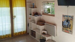 Ein Zimmer mit Holzboden, einem Glastisch, einem Ventilator, Regalen mit diversen Gegenständen, einem Fenster mit Blick und einem Fernseher an der Wand.