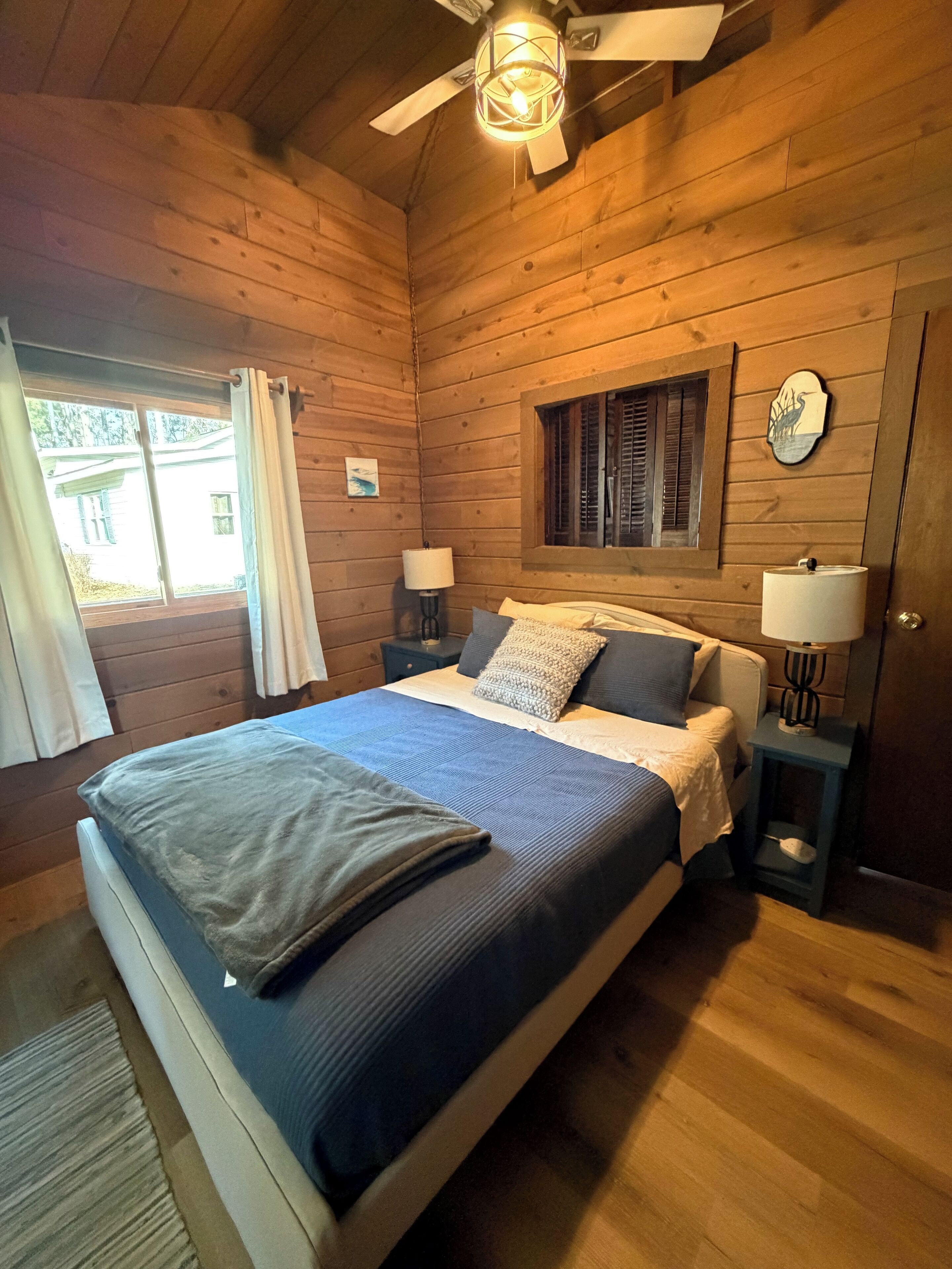 Une chambre dans une cabane en bois, avec un lit, deux tables de chevet avec des lampes, une fenêtre avec des rideaux et un ventilateur de plafond.