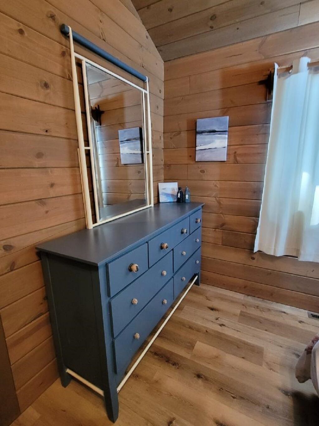A wooden-paneled room with a dark blue dresser, a mirror, and a small table with bottles.