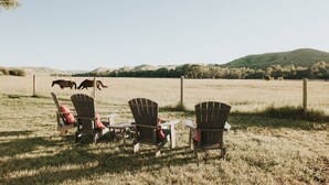 Three Adirondack chairs with red cushions in a grassy field with a view of hills and a fence in the background. - Big Sky Escape: Spacious Home on Working Ranch (Reed Point)