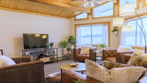 A cozy living room with leather sofas, a flat-screen TV, wooden flooring, and large windows offering a view of the outdoors. - Big Sky Escape: Spacious Home on Working Ranch (Reed Point)