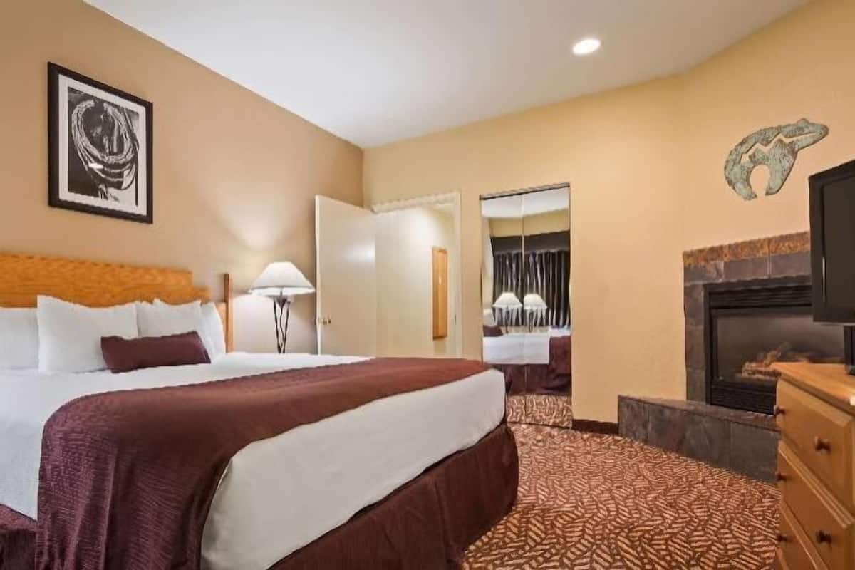 A hotel room with a large bed, a television, a fireplace, and a dining area visible through an open door.