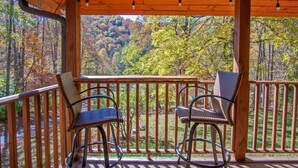 A wooden deck with two wicker chairs and a table, overlooking a forested area with autumn foliage.
