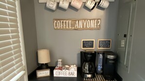 A coffee bar with a sign that reads "COFFEE, ANYONE?" and a sign that says "Coffee Bar". There are coffee mugs hanging on the wall and a coffee maker on the counter.