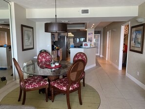A dining area with a glass table, four chairs with star patterns, a hanging light fixture, and a kitchen in the background.