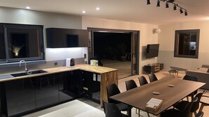 A modern kitchen with a built-in island, a dining area with a long table and chairs, and a TV mounted on the wall.