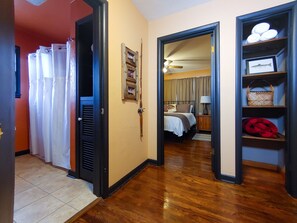 Een hotelkamer met een houten vloer, een bed met witte lakens, een nachtkastje met een lamp en een plank met handdoeken en een mand.