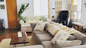 Ein Wohnzimmer mit einer Couch, einem Hocker und einem Couchtisch. Im Raum befindet sich eine Tür, eine Pflanze und ein Sessel.