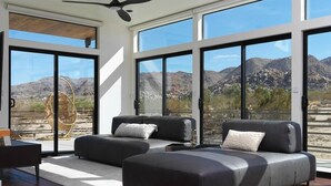 A modern living room with a large window, a ceiling fan, and a view of mountains. - Panoramic Heaven - Luxury Property Pool & Spa (Joshua Tree)