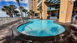 A swimming pool with a curved design, surrounded by lounge chairs and palm trees, adjacent to a building with a sign indicating restrooms. - Low Floor, beautiful ocean views, seasonal beach chair service. No elevator wait (Panama City Beach)