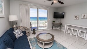A living room with a sofa, a coffee table, a TV, and a balcony with a view of the ocean. - Low Floor, beautiful ocean views, seasonal beach chair service. No elevator wait (Panama City Beach)