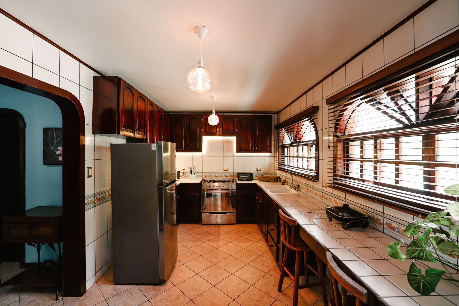 A compact kitchen with a refrigerator, stove, and sink. There is a window with blinds and a hanging light fixture.