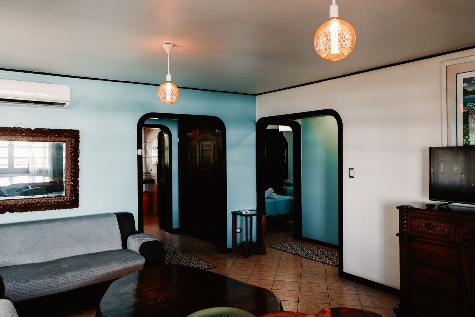 A living room with a sofa, a coffee table, and a TV. There are two hanging lights and a mirror on the wall.