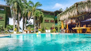 A pool area with a clear blue pool, palm trees, and a building with a green exterior and red window frames.
