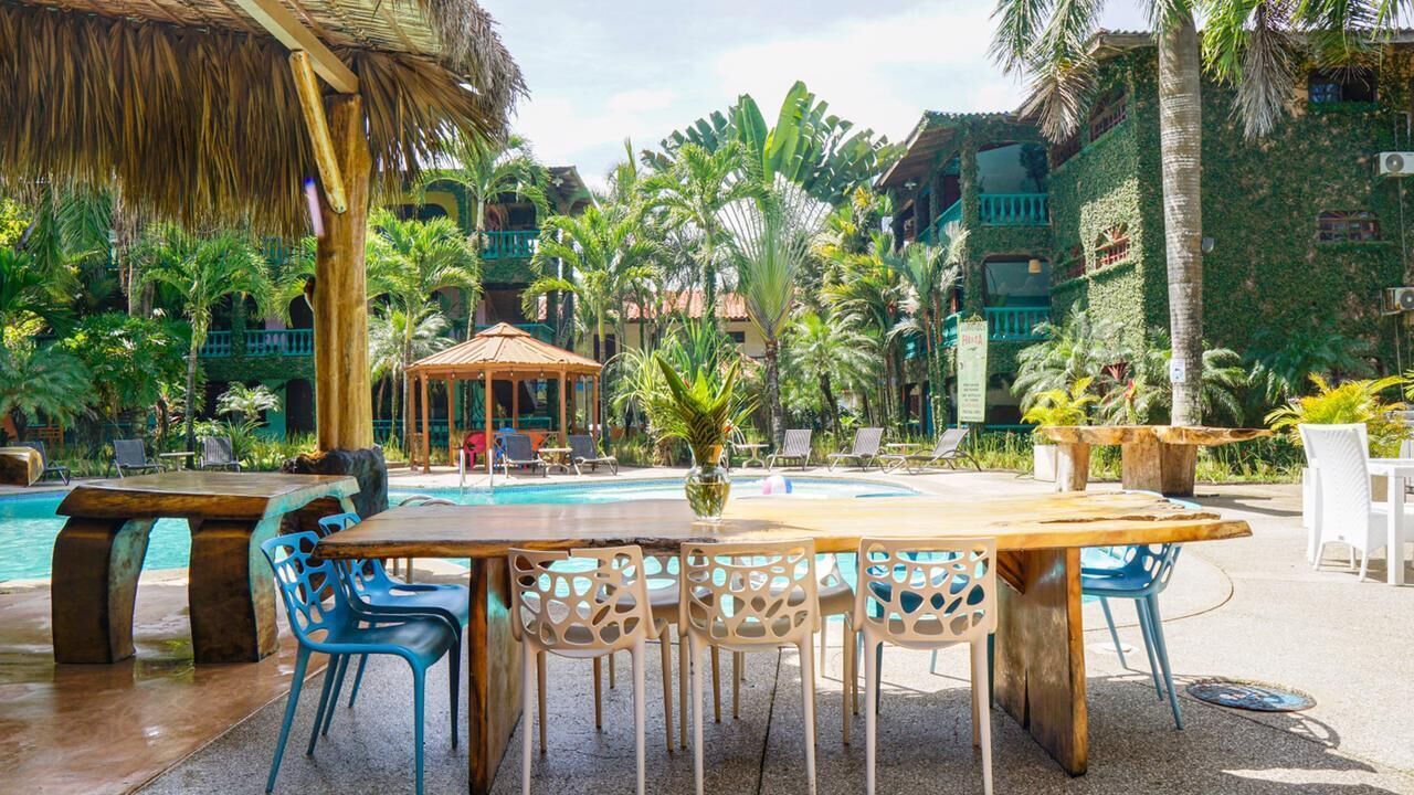A poolside area with a wooden table and blue chairs, surrounded by palm trees and a thatched roof structure.
