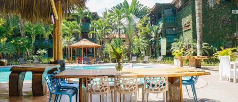 A poolside area with a wooden table and blue chairs, surrounded by palm trees and a thatched roof structure.