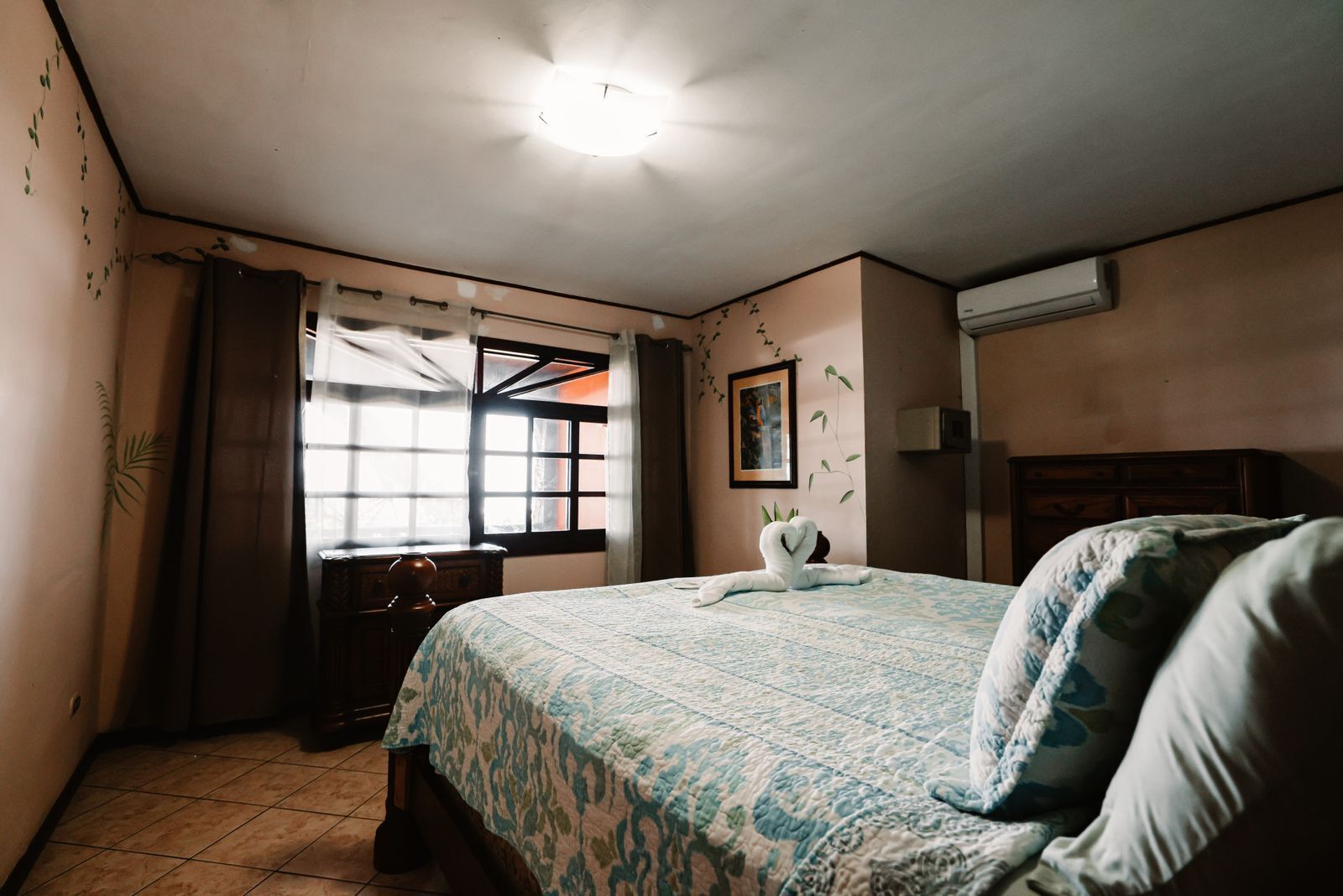 Un dormitorio con cama, ventana con cortinas, aire acondicionado de pared y un cuadro enmarcado.