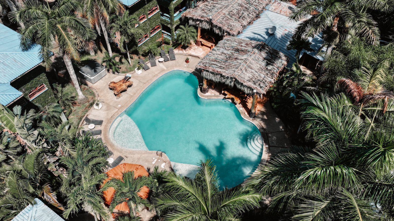 Vista aérea de un resort con una piscina central rodeada de palmeras y estructuras de techo de paja.