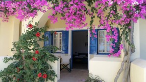 A house with a porch covered in pink flowers, blue shutters, and a visible doorway leading inside. - Santorious Cavehouse (Santorini)