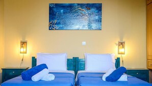 A hotel room with two beds, each with a blue mattress and white pillows, and a painting above the beds. - Santorious Cavehouse (Santorini)