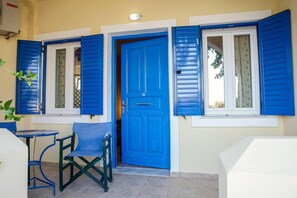 A room with a blue door, two windows with blue shutters, a small table, and a chair. - Santorious Cavehouse (Santorini)