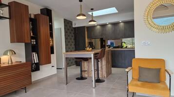 A modern kitchen with dark cabinetry, a wooden island, and a bar area. There is a yellow chair and a round mirror with a sunburst design on the wall.