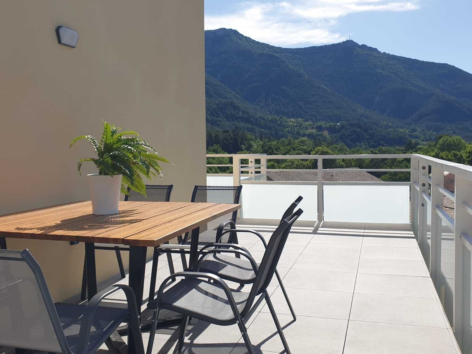 Een balkon met een houten tafel en stoelen, een plant in een pot en uitzicht op bergen.
