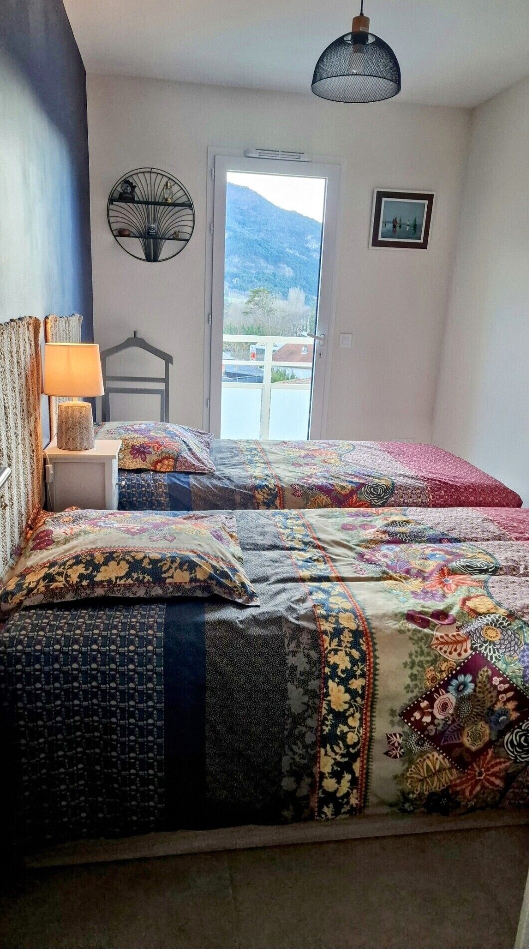 A bedroom with a large bed, a bedside table with a lamp, a hanging chair, and a view of a mountain through a window.