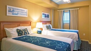 Two beds with blue and white patterned bedspreads, a wall-mounted quote, and a window with beige curtains.