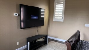 Quarto com televisão de tela plana na parede, um banco de madeira, uma janela com persianas e um edredom estampado.