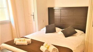 A neatly made bed with a dark headboard, white bedding, and two pillows. There is a bench at the foot of the bed and a door to the left.