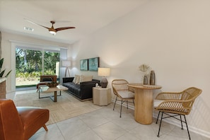 A modern living room with a sofa, coffee table, and chairs. There is a ceiling fan, a lamp, and a view of the outdoors.