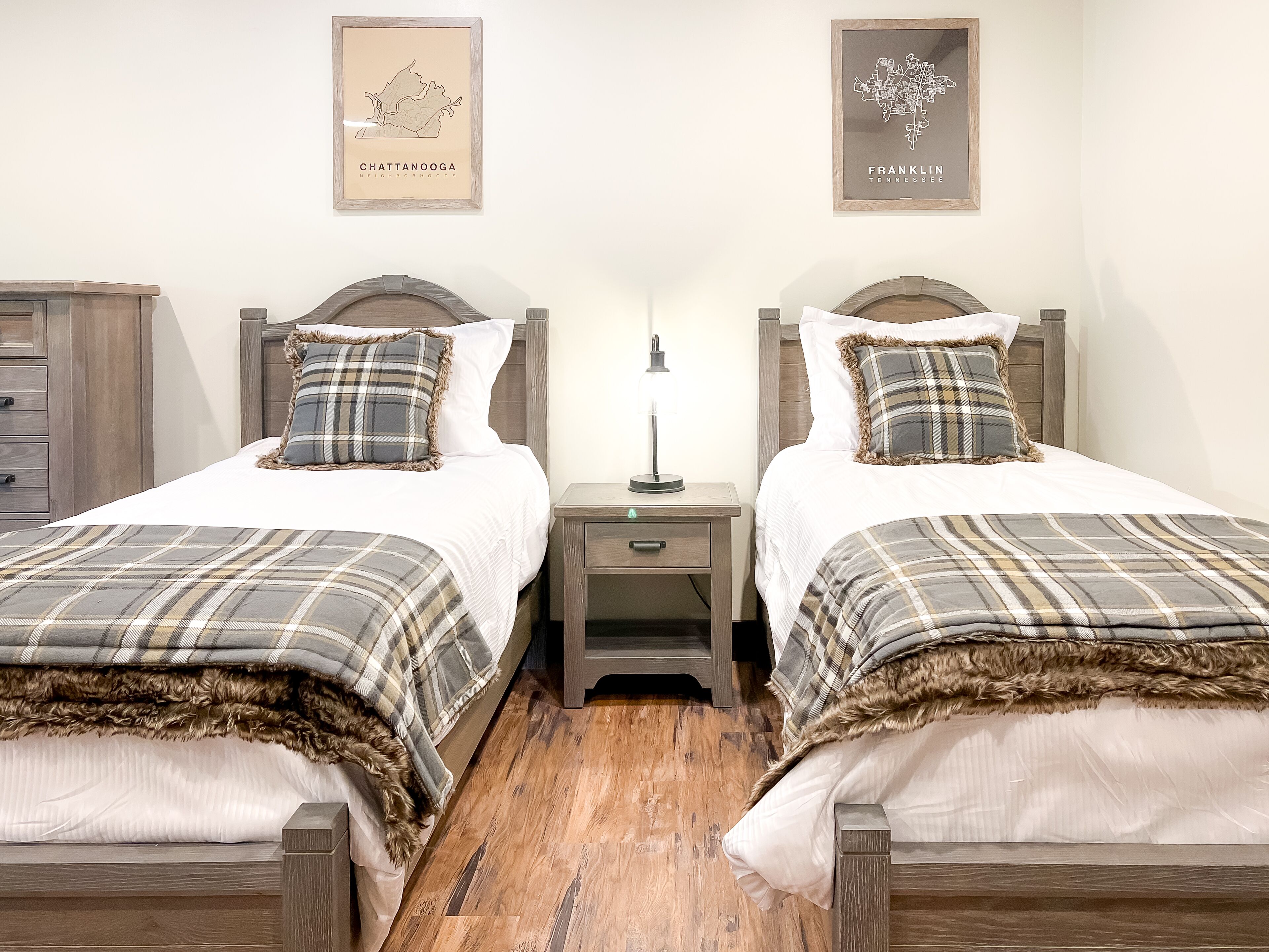 Une chambre double avec deux lits, chacun recouvert d’une couverture à carreaux et agrémenté d’oreillers assortis. Les murs sont ornés de cartes encadrées, et la chambre comprend une table de chevet avec une lampe, ainsi qu’un plancher en bois.