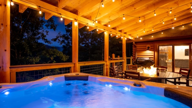 A hot tub with a wooden deck and string lights, surrounded by wooden furniture and a fire pit. - Luxurious Cabin / Theater / Game Room / Hot Tub / Large Deck / Panoramic View (Sevierville)