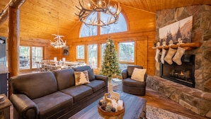 A cozy cabin interior with a fireplace, a Christmas tree, and a large window overlooking a forest. - Luxurious Cabin / Theater / Game Room / Hot Tub / Large Deck / Panoramic View (Sevierville)