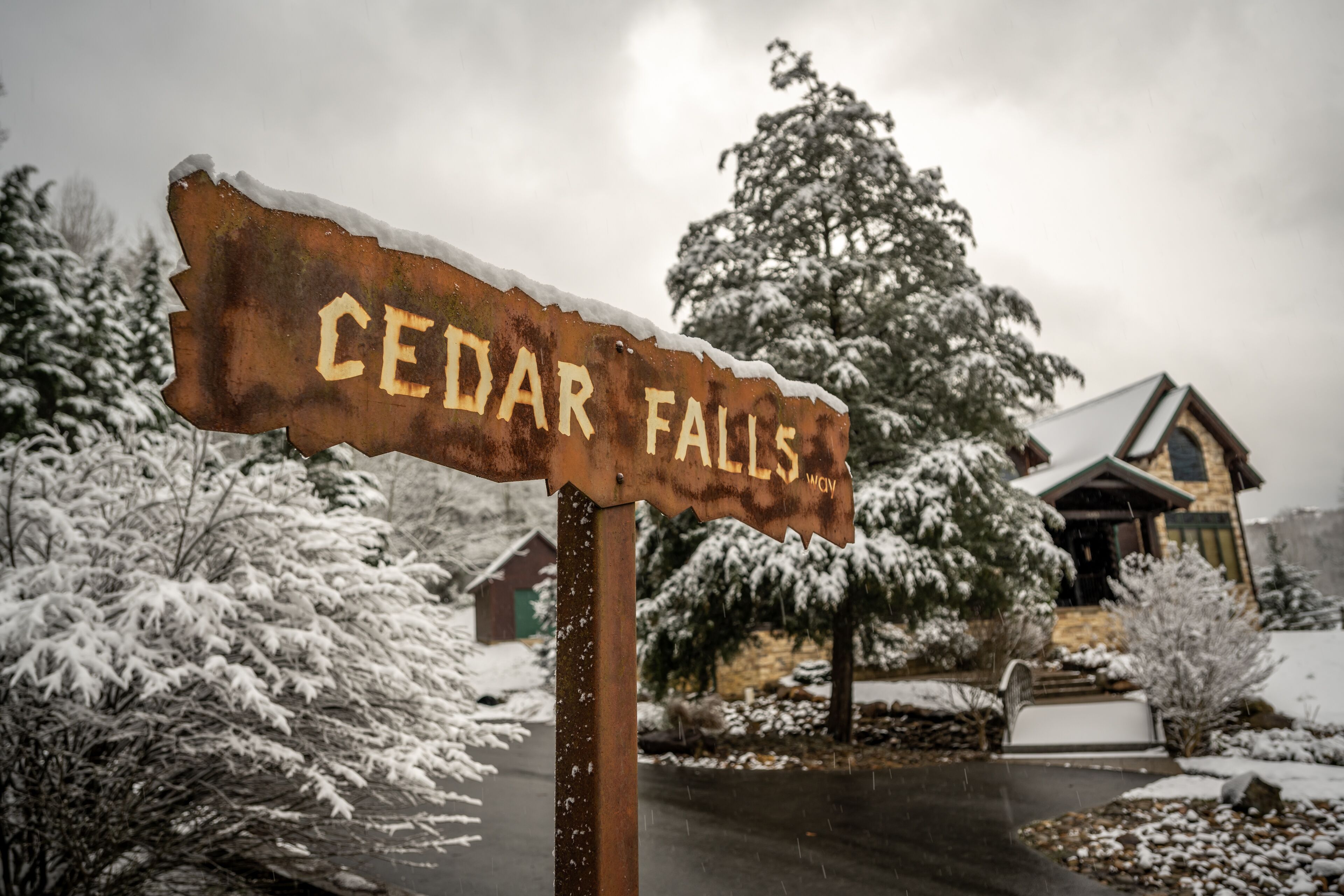 Ein verwittertes Schild mit der Aufschrift „CEDAR FALLS“, im Hintergrund verschneite Bäume und ein Haus.