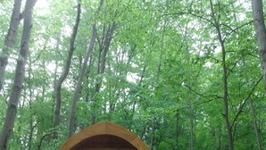 A wooden cabin with a large window and an arched roof, surrounded by a dense forest. - Lookout Camping Pod (Freeville)
