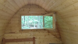 A cozy cabin room with a wooden ceiling, a bed with white bedding, a blue chair, and a window offering a view of the forest. - Lookout Camping Pod (Freeville)