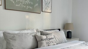 A neatly made bed with striped bedding and a 'Welcome Home' pillow, a bedside lamp, and two framed wall art pieces.