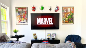 A living room with a flat-screen TV, two framed comic books, a small table with a plant, and a sofa.