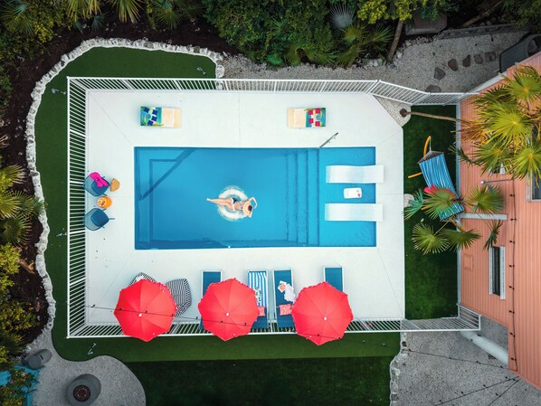 Luftaufnahme eines Schwimmbeckens mit Liegestühlen, Sonnenschirmen und einer Person, die schwimmt.